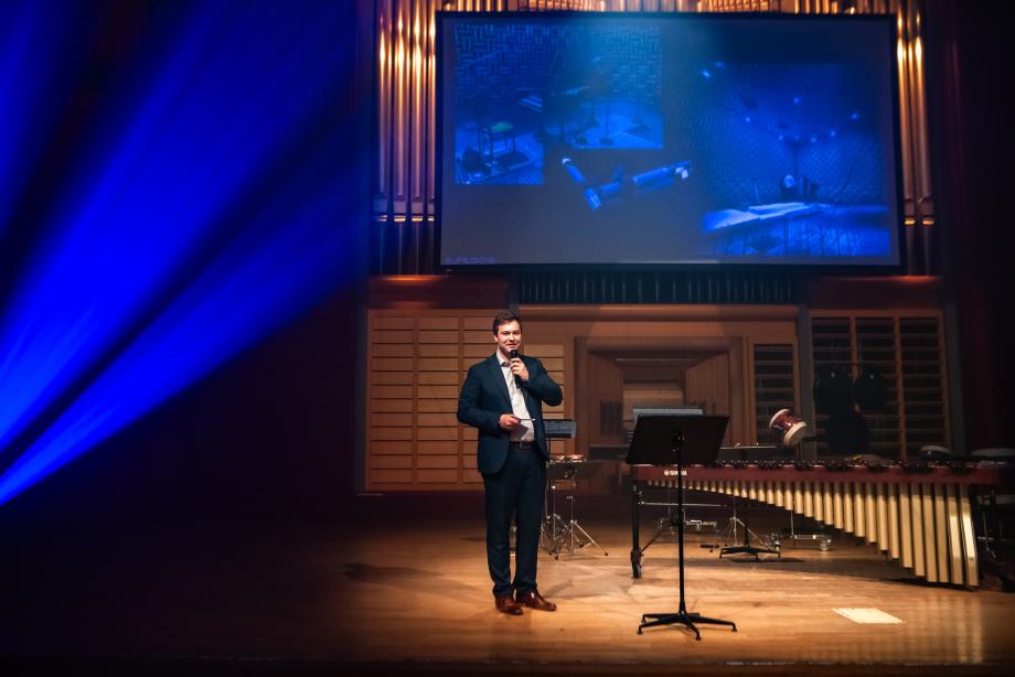 Arne Morgner (Leitung Tonstudio und Veranstaltungstechnik) steht sprechend mit Mikrofon auf der Bühne neben Percussioninstumenten. Hinter ihm ist eine große Leinwand mit Bildern von Instrumenten in einer Art Tonstudio zu sehen.