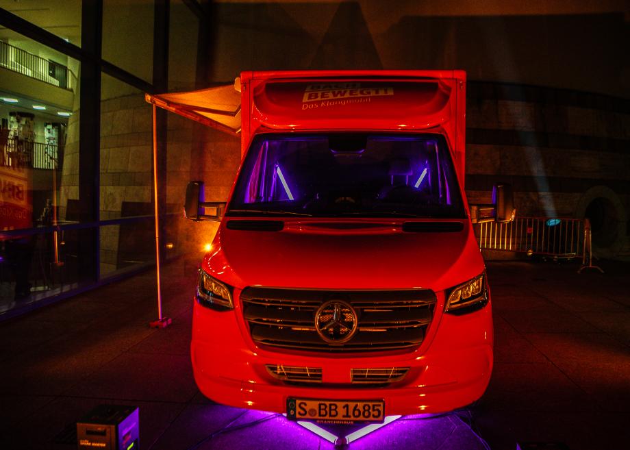 Frontansicht eines roten Mercedestransporters bei Nacht, mit violettem Licht im Innenraum und unter dem Fahrzeug, vor der HMDK.