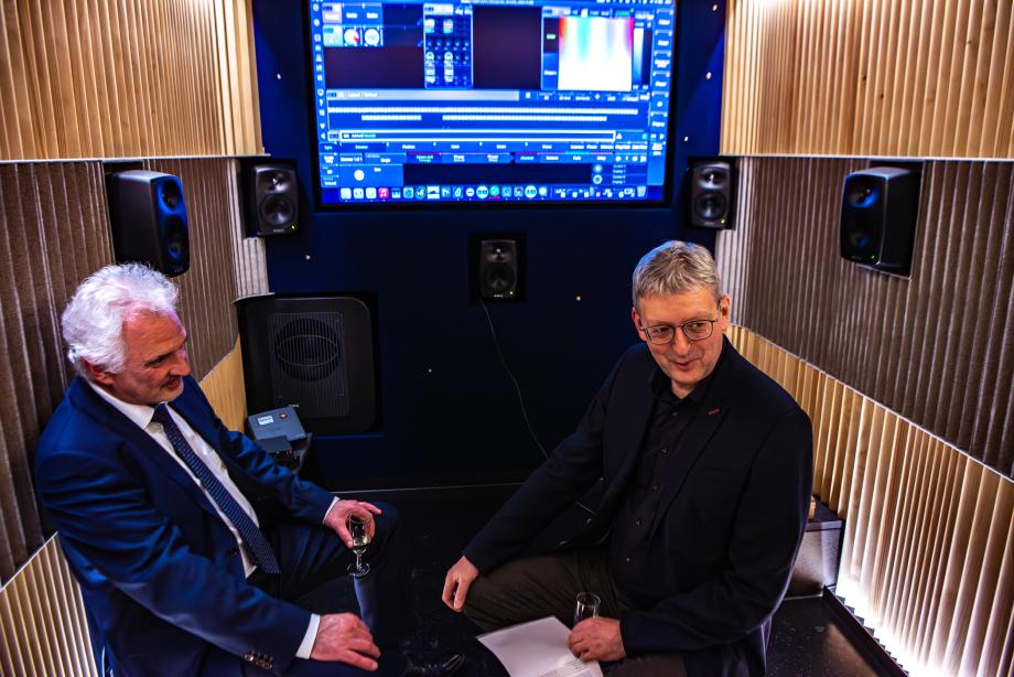 Hans-Christoph Rademann (Akademieleiter Internationale Bachakademie Stuttgart) und Axel Köhler (Rektor der Staatlichen Hochschule für Musik und Darstellende Kunst) stehen im Innenraum des Klangmobils (kleiner Konzertsaal) mit gelb beleuchteten, vertikal geriffelten Wänden, schwarzem Boden und großer Leinwand mit Computerbildschirm.