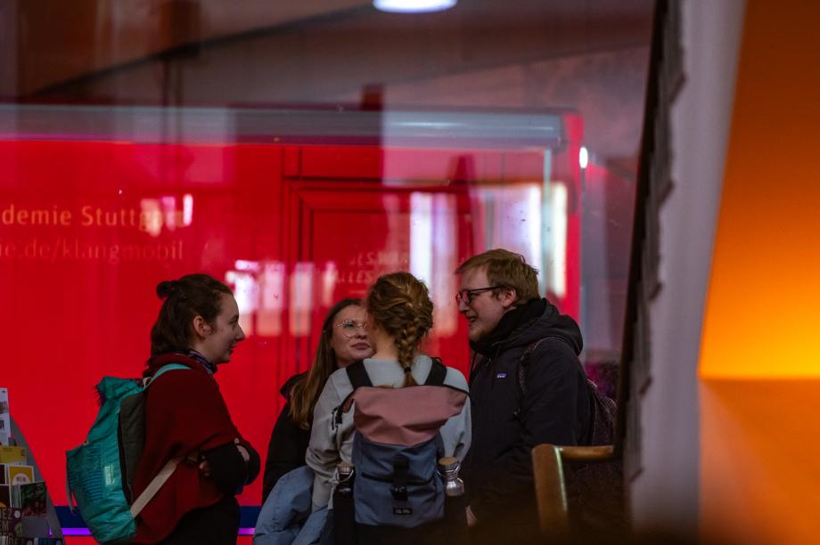 Einige junge Menschen vor den Glasfenstern der Musikhochschule. Im Hintergrund sieht man den roten Mercedestransporters bei Nacht.