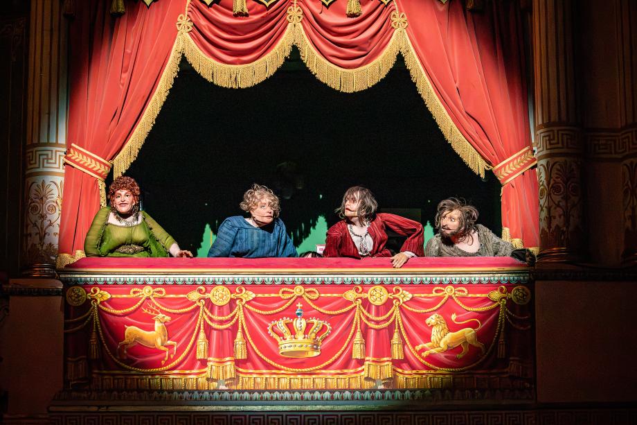 Vier Puppen, in unterschiedlichen Kostümen, schauen aus der Loge im Wilhelma Theater in den Saal. Die jeweiligen Gesichter der puppen sind eher grob gearbeitet. Alle vier werden von einem Scheinwerferspot angestrahlt.b