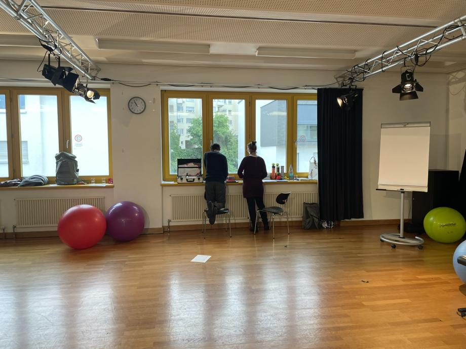 Zwei Personen stehen am Fenster und machen sich gemeinsam Notizen zu einer Aufgabe. Links neben ihnen befindet sich ein Koffer mit Moderationsmaterialien und auf der anderen Seite ein Flipchartständer mit Papier. Im Raum verteilt sieht man drei Gymnastikbälle.
