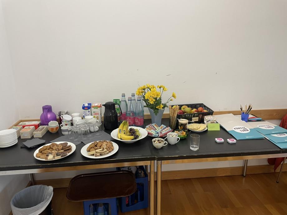 Zu sehen ist das Kaffeebuffet mit Keksen, Getränken und Blumen im Seminarraum. Auf der rechten Seite des Tisches liegen Teilnehmer-Mappen mit dem Logo der HMDK und der Aufschrift „Künste im Turm“.