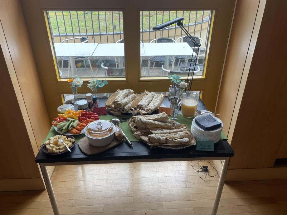 Man sieht einen Tisch mit Mittagsbuffet vor einem großen Fenster. Das Buffet besteht aus einem großen Rohkostteller, Käsewürfeln, Suppe und gefüllten, gerollten Naanbroten.
