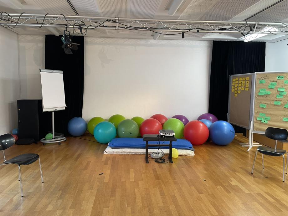 Ein Raum, an dessen Rückwand 14 Gymnastikbälle aufgereiht sind, davor liegen Gymnastikmatten für Körper- und Stimmübungen. Rechts stehen zwei Pinnwände mit gelben und grünen Post-its, auf denen Seminarinhalte stehen. Links steht ein Flipchartständer mit Papier. Ein Beamer steht in der Mitte des Raumes und ist auf die Rückwand ausgerichtet.