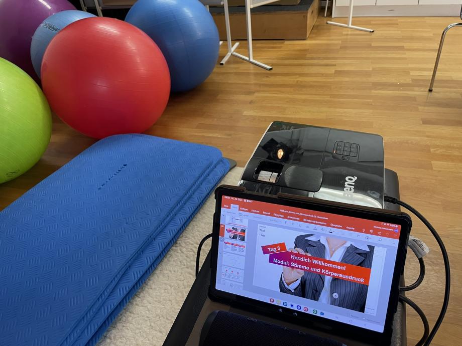 Nahaufnahme eines Laptopbildschirmes im Präsentationsmodus. Darauf ist eine Person zu sehen, die ein Schild in der Hand hält mit der Aufschrift „Tag 3 – Herzlich willkommen. Modul: Stimme und Körperausdruck“. Im Hintergrund sind Gymnastikbälle - und Matten.