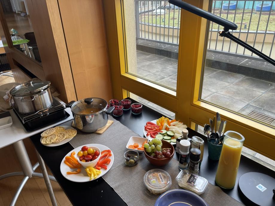 Man sieht einen Tisch mit Mittagsbuffet vor einem großen Fenster. Das Buffet besteht aus Linsensuppe, frischem Obst und Gemüse und einem rote Bete Salat.
