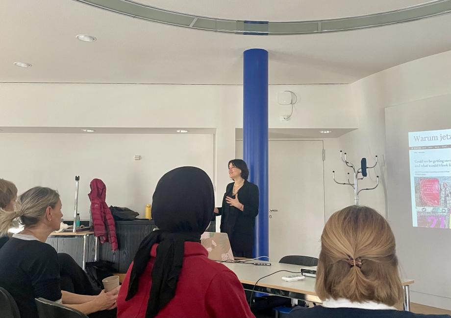 Die Referentin steht vor einer blauen Säule und gestikuliert vor den Zuhörenden.