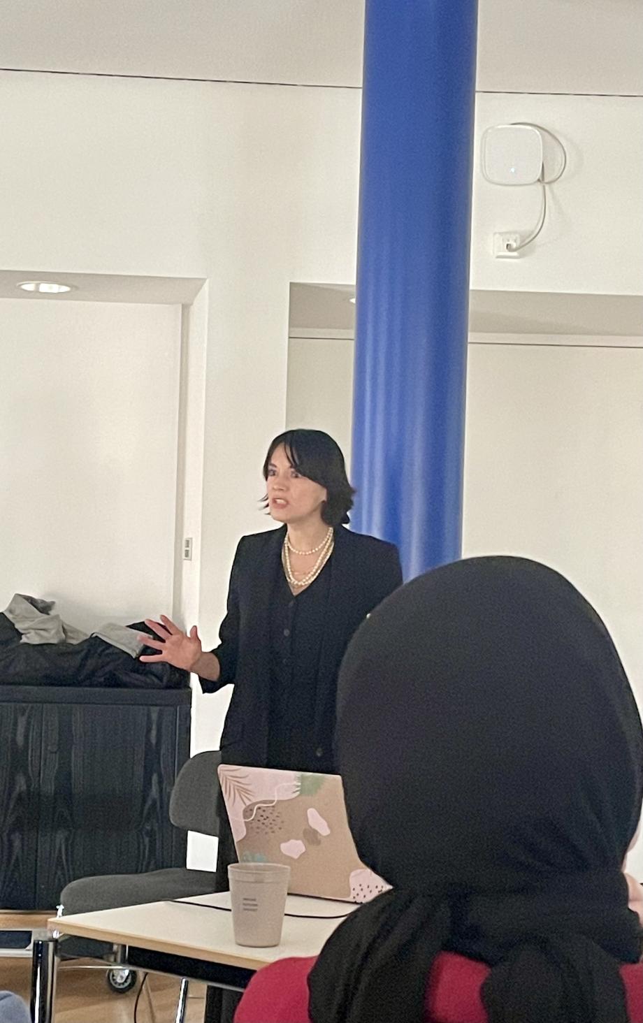 Die Referentin steht vor einer blauen Säule und gestikuliert vor den Zuhörenden.