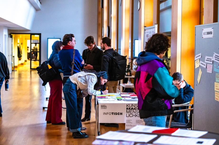 Studentische Teilnehmende in der Wandelhalle beim Infotisch im Gespräch und Notizen machend.