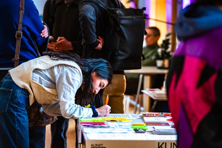 Studentische Teilnehmende in der Wandelhalle beim Infotisch Notizen machend.
