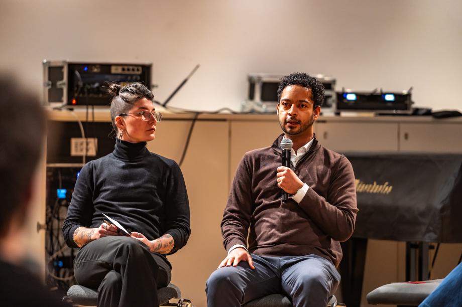 Leonid Mauch spricht in ein Handmikrofon, Celin Klotz sitzt neben ihm und hört ihm  aufmerksam zu.