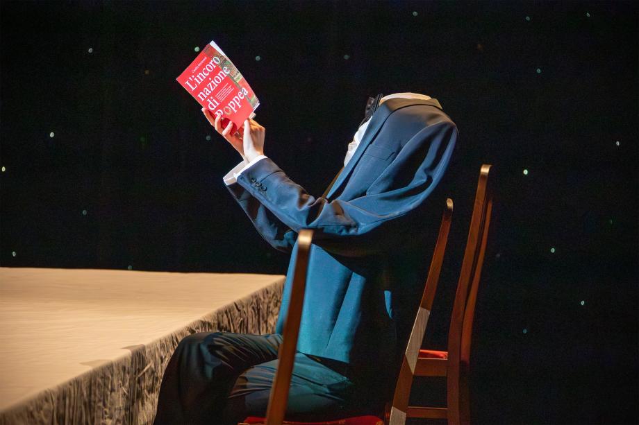 Ein kopfloser Mann liest an einem Tisch sitzend das Programmheft von "Poppea"