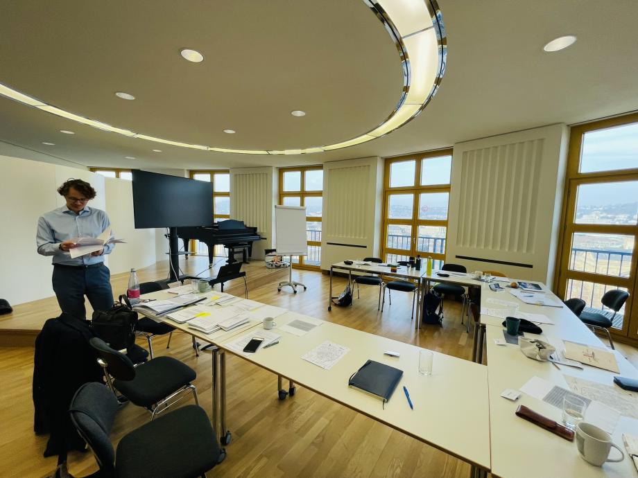 Ein großer Raum mit Ausblick auf Stuttgart. Die Tische stehen in U-Form, darauf liegen Arbeitsmaterialien, Blätter und Kaffeetassen. Links im Bild steht ein Dozent, der seine Unterlagen durchblättert.