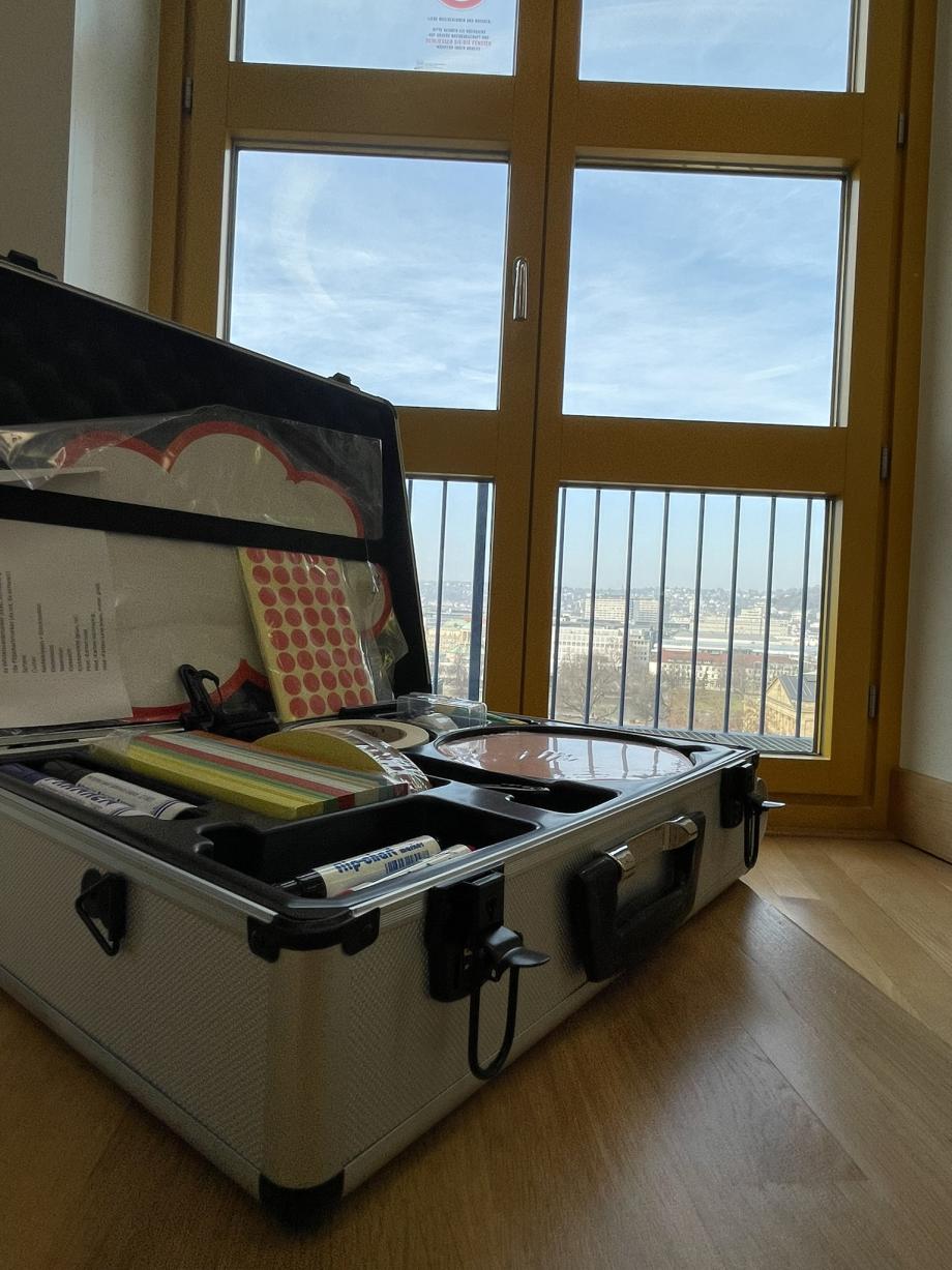 Ein geöffneter Alukoffer (Moderationskoffer) steht auf einem Podest vor dem Fenster, durch das man Ausblick auf Stuttgart hat. Im Koffer sieht man Materialien wie Flipchartstifte, Moderationskarten und Klebepunkte.