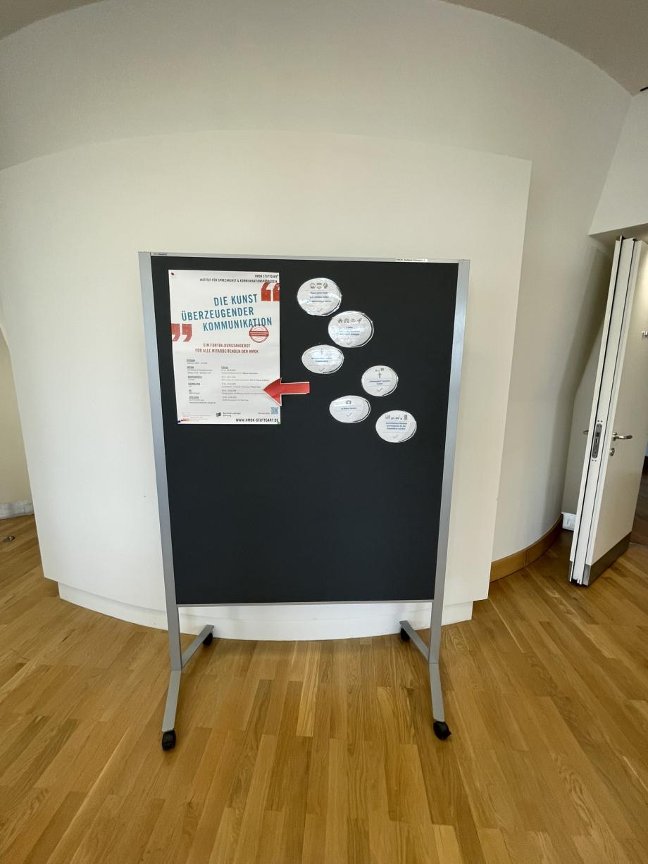 Eine mobile Pinnwand mit Rollen vor einer halbrunden weißen Wand. An der Pinnwand hängt das Plakat mit dem Slogan „Die Kunst überzeugender Kommunikation“ und den Namen und Terminen der einzelnen Module. Rechts neben dem Plakat hängen sechs runde, laminierte Karten, auf denen die Fortschritte und Erkenntnisse der Fortbildungsgruppe stehen.