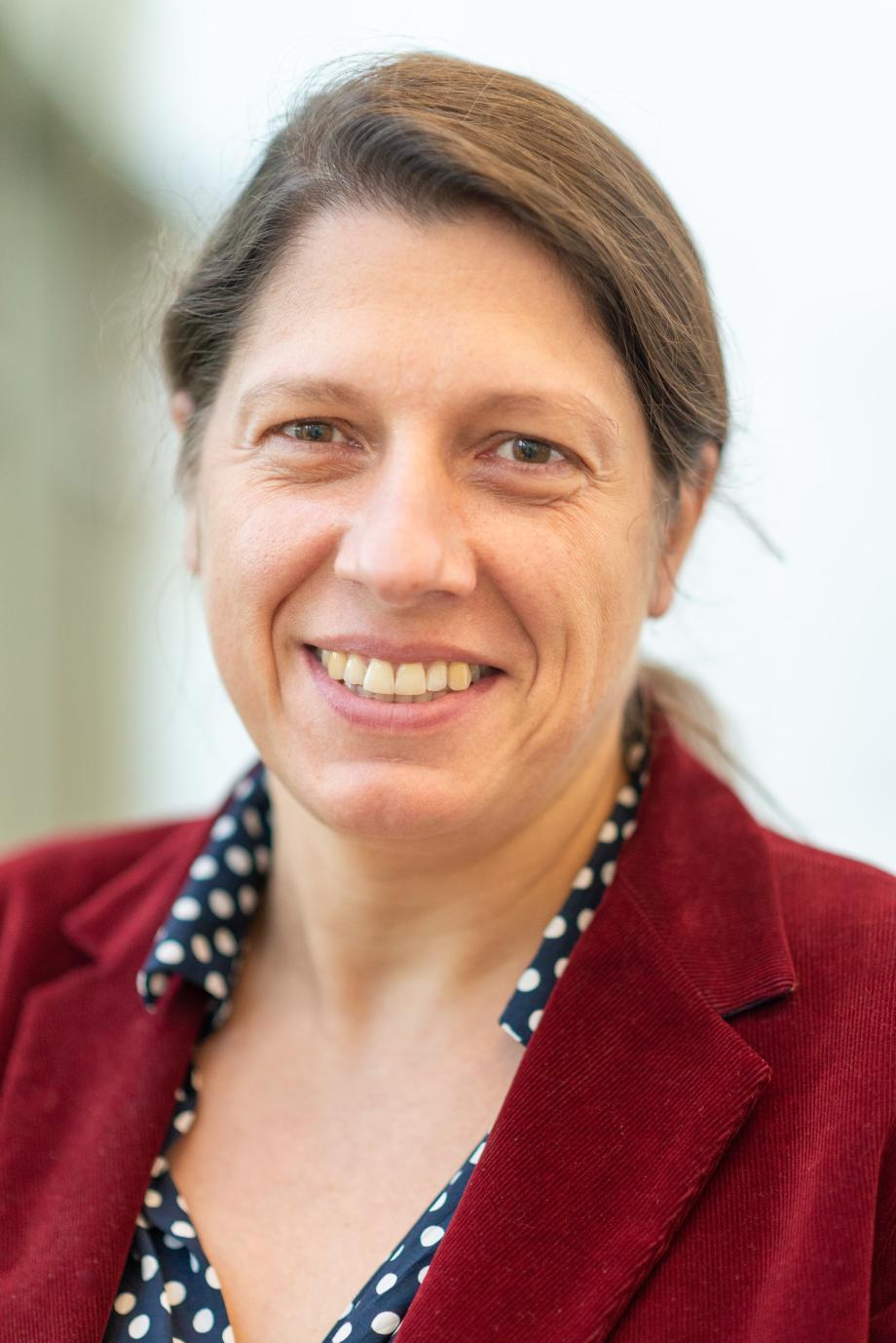 Portrait von Prof.in Dr. Constanze Spieß. Sie hat dunkelblonde Haare zu einem Zopf gebunden, nicht geschminkt, und schaut lächelnd in die Kamera. Sie trägt einen roten Cord-Blazer und eine blaue Bluse mit weißen großen Punkten.