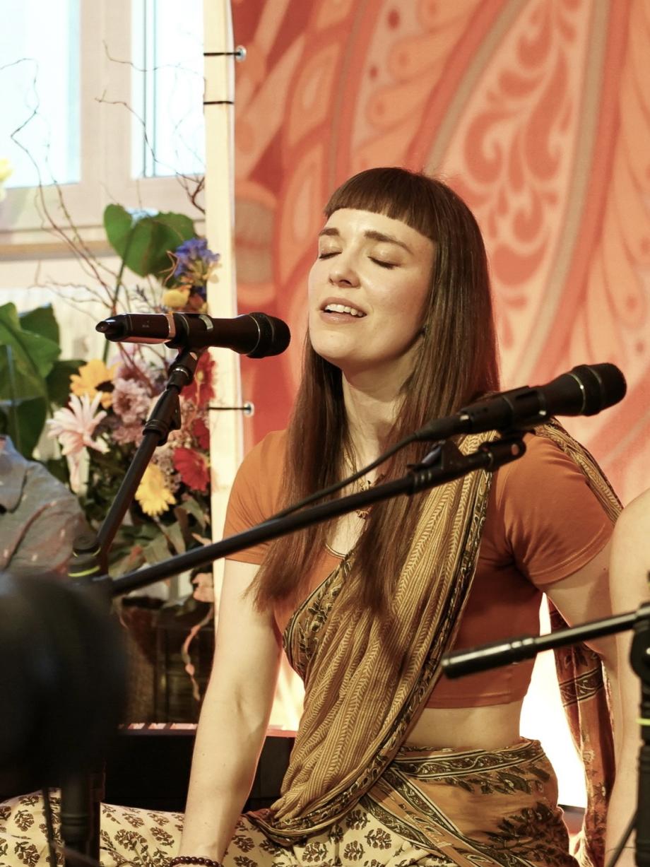 Fabienne Fleischmann trägt indische traditionelle Kleidung in Erdtönen und sitzt auf dem Boden. Sie hat lange braune offene Haare, einen kurzen Pony und ihre Augen sind geschlossen und sie singt in ein Mikrophone.