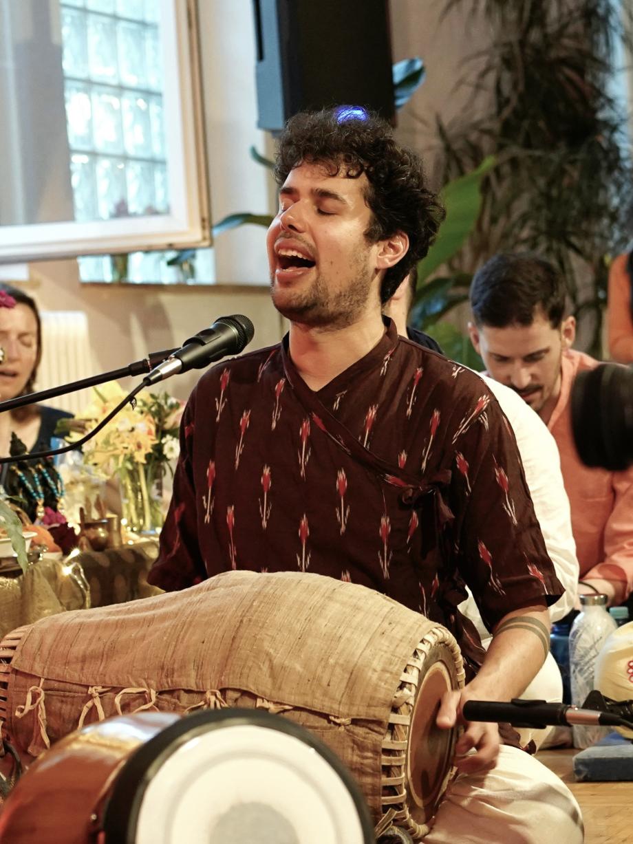 Ein Student sitzt im Schneidersitz auf dem Boden. Auf seinem Schoß hat er eine Trommel auf der er spielt. Seine Augen sind geschlossen und er singt in ein Mikrophone.