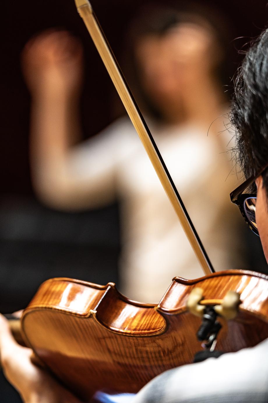 Blick über die Schulter eines Violinenspielers. Im Hintergrund ist verschwommen ein Dirigent zu sehen.