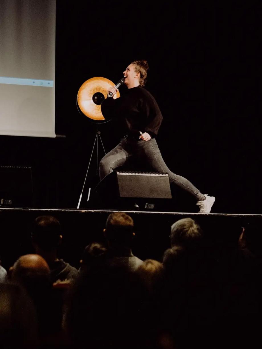 Lena Stokoff läuft auf der Bühne. Vor der Bühne sind Menschen von hinten zu sehen, die Lena Stokoff beobachten. Lena Stokoff trägt eine enge graue Jeans mit weißen Turnschuhen und einem dunklen Pullover. Sie ist von der Seite zu sehen und schaut nach oben. Sie hat ein Mikrophone in der Hand und spricht und lacht.