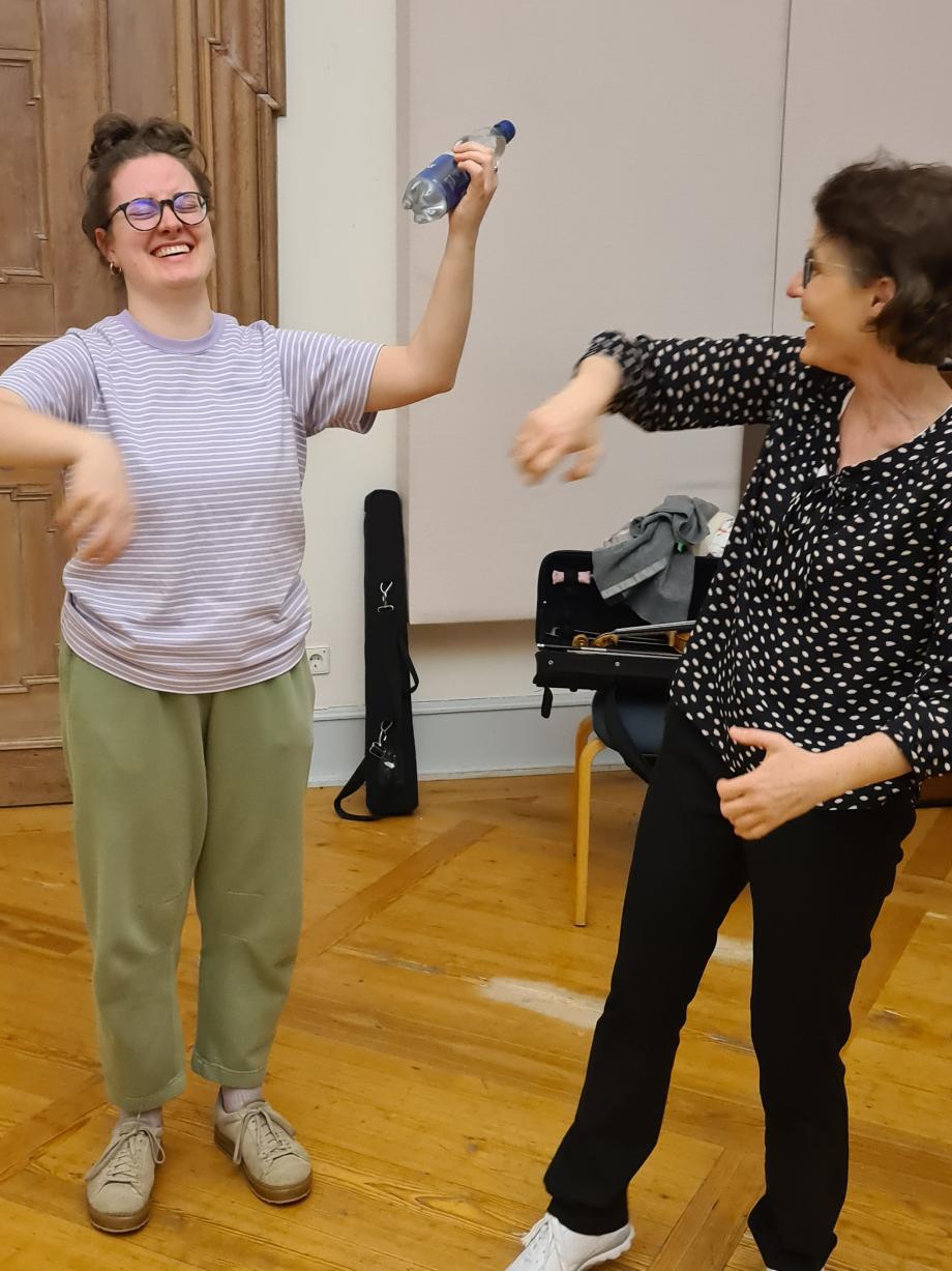 Eine Dozentin übt mit einer Studierenden Bewegungstechniken für das Violinspiel. Sie haben beide viel Spaß dabei und lachen. Die Übung ist ohne Instrument sondern mit einer kleinen Wasserflasche anstelle des Griffbretts.