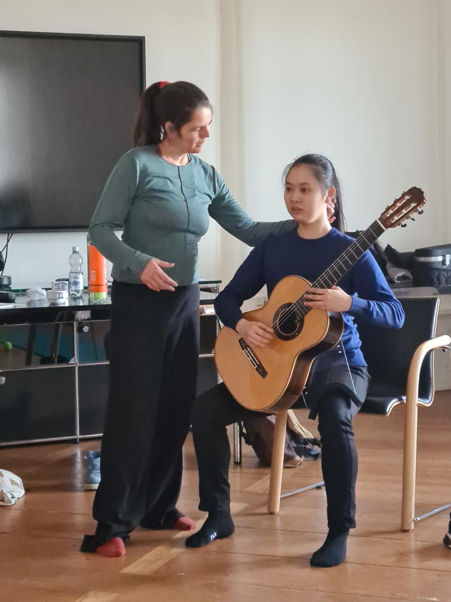 Eine Studentin mit Gitarre übt barfuß auf einem Stuhl sitzend. Die Dozentin korrigiert mit der Hand an ihrem Nacken die Haltung.