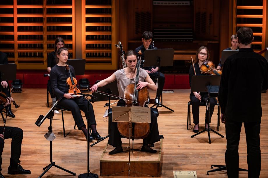 Preisträgerin des Cellowettbewerbs 2022 spielt beim Preisträgerkonzert auf der Bühne des Konzertsaals. Sie sitzt auf einem kleinen Podest und hinter ihr mehrere Streicher und Streicherinnen.
