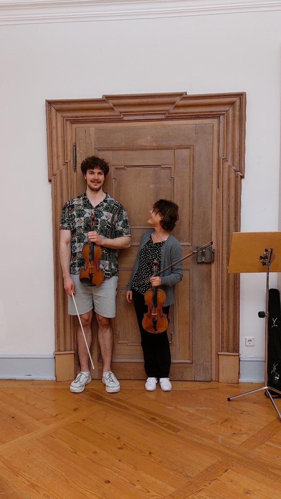 Violinstudent und Dozentin stehen neben einander. Die Dozentin ist sichtbar kleiner als ihr Schüler. Sie schaut amüsiert zu ihm auf.