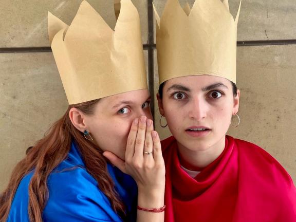 Eine Studierende mit Krone flüstert einer anderen bekronten Studierenden etwas zu. 