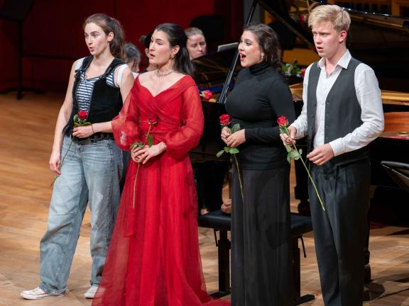 Ein Opernquartett aus Studierenden musiziert auf dem Konzertsaalpodium vor einem Flügel. Alle vier Studierende tragen eine rote Rose in ihrer hand.