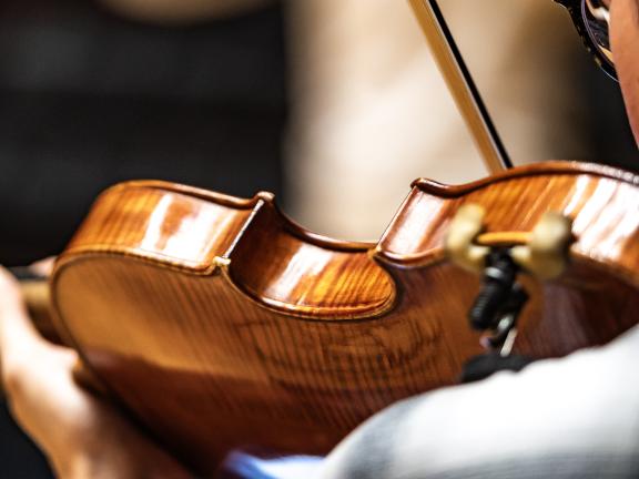 Blick über die Schulter eines Violinenspielers. Im Hintergrund ist verschwommen ein Dirigent zu sehen.