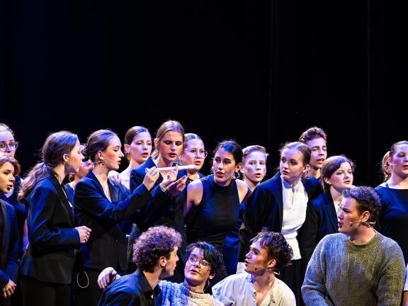 Ein Schüler*innenchor steht auf der Bühne, alle blicken auf eine Flöte in der Mitte