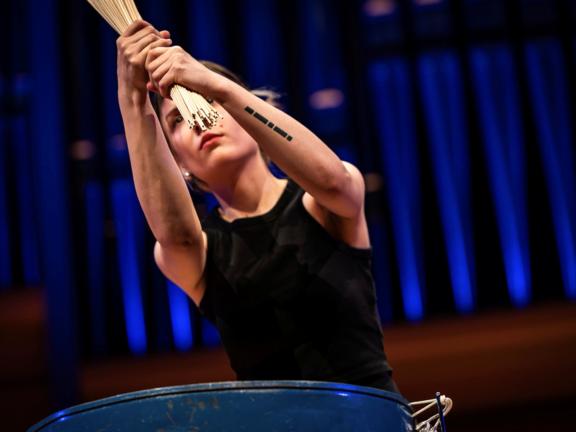 Die Schlagzeugerin Tamara Kurkiewicz hält mit beiden Händen Holzstäbe vor sich. Im Hintergrund sieht man das blauerleuchtete Orgelprospekt.