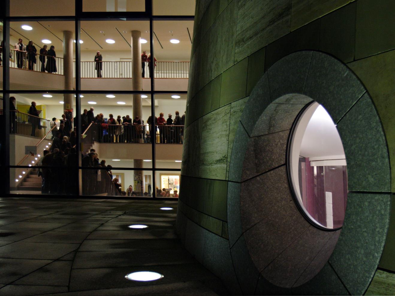 Die Außenansicht des Turms bei Nacht. Im Hintergrund sind Menschen durch die Glasfassade im den Hochschulfoyers zu sehen.r zu sehen. 
