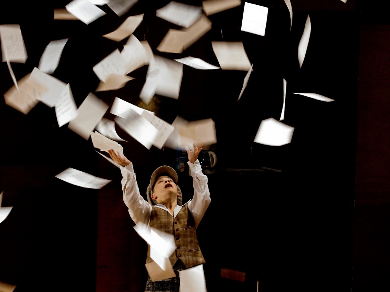 Eine Person hinter einem Holztisch wirft weiße Blätter in die Luft