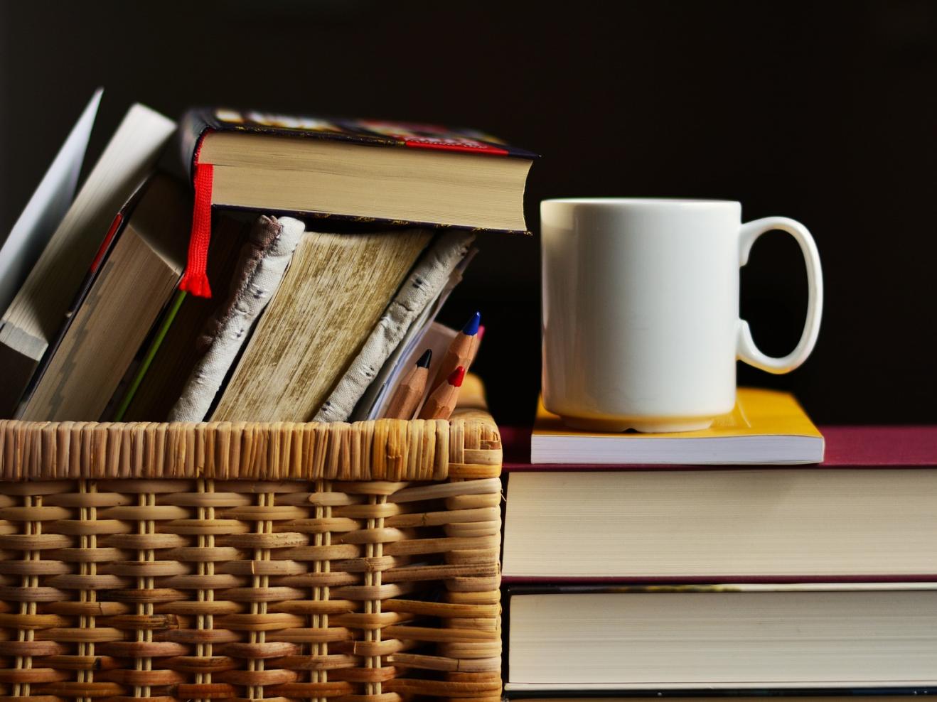 Bücher in einem Korb, daneben steht eine weiße Tasse auf zwei dicken Büchern.