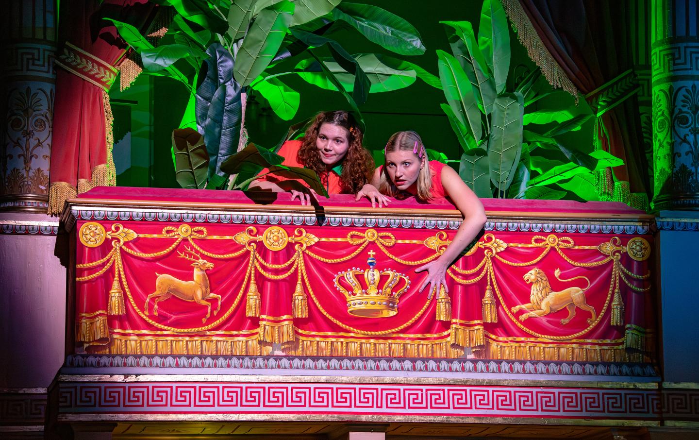 Zwei Schauspielstudierende schauen aus einer mit Palmen dekorierten Loge des Wilhelma Theaters haus. Die eine eher ungläubig und erschrocken. Die andere interessiert und vorfreudig