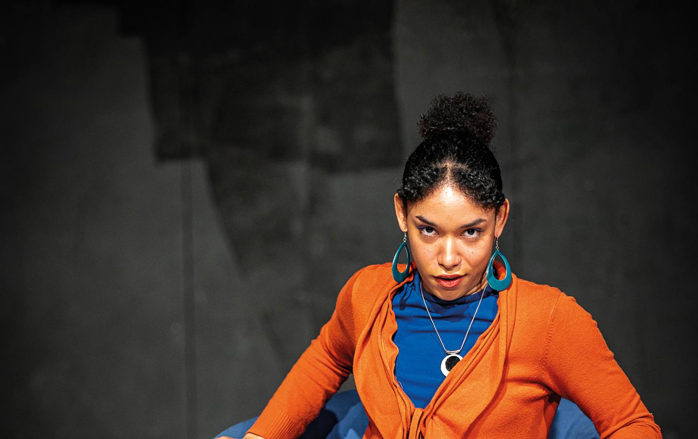 Schauspielstudentin mit aufgeschlagenem großen Buch auf dem Schoß sitz in einem Sessel und schaut intensiv in die Kamera.