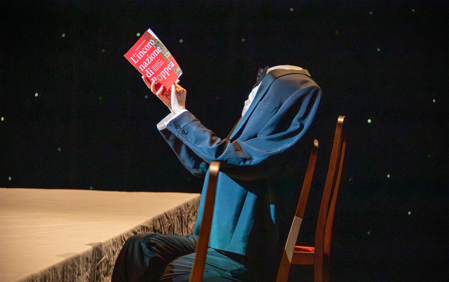 Ein kopfloser Mann liest an einem Tisch sitzend das Programmheft von "Poppea"